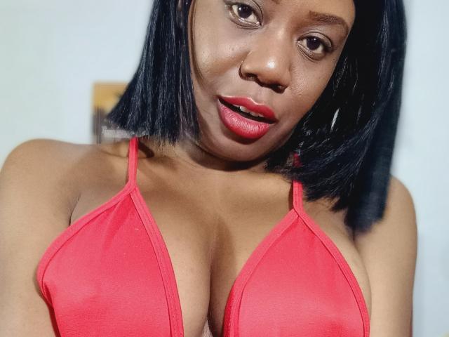 Karhinna with dark skin and black shoulder-length hair, wearing a red bikini top, showing her big, round tits and red lipstick. She has a slight, sexy smile.