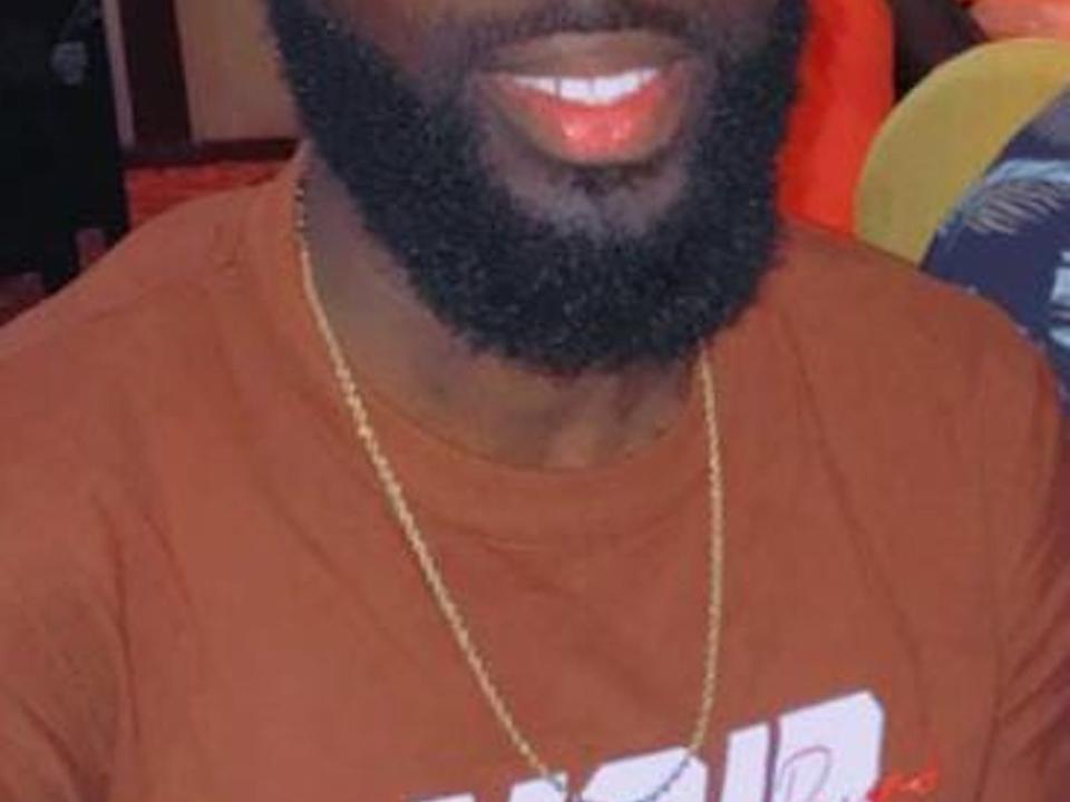 La barba scura e riccia di RebellKing e le sue labbra rosse lucide sono visibili. Indossa una camicia marrone e una catena d'oro, e la parte superiore del suo corpo è inquadrata..