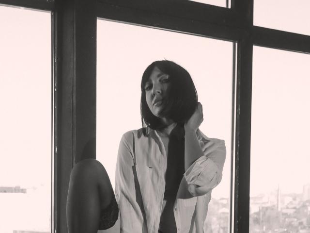 Black-and-white photo of MissGulia with short dark hair, wearing an unbuttoned shirt, exposing her dark lace bra and thigh, leaning against a window with her hand on her neck.