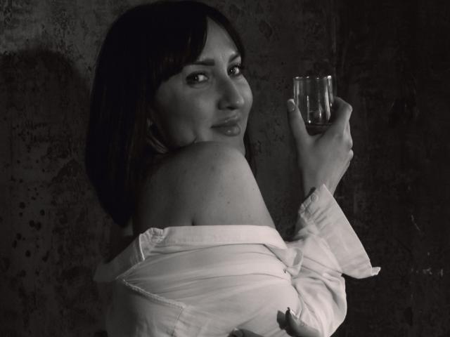 MissGulia with dark hair, looking back, holding a glass, wearing an off-shoulder white shirt, exposing her shoulder and upper back, in a black-and-white photo.