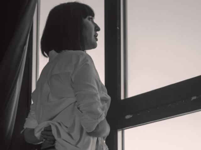 Black and white photo of MissGulia with short dark hair, wearing an unbuttoned shirt, standing in profile by a window, hands behind her back, looking outside.