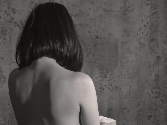 Black-and-white photo of MissGulia with shoulder-length dark hair, back to camera, topless, showing bare shoulders and upper back, with a slightly textured wall behind her.