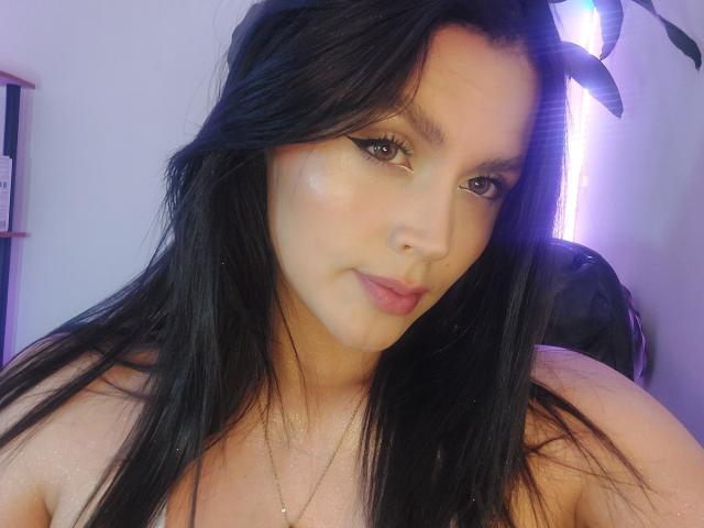 LinaStars with long black hair, brown eyes, and pink lips, wearing a necklace, looking at the camera with a slight smile. Her face is slightly shiny.