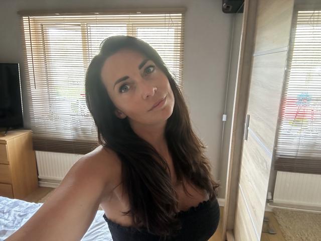 BiancaGlam with long brown hair, wearing a black strapless top, showing her boobs, taking a selfie in a sunlit room, standing in front of a mirror.