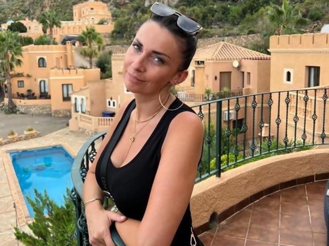 BiancaGlam with dark hair in a bun, wearing a black tank top, hoop earrings, and sunglasses on her head, standing by a pool with a curved balcony.