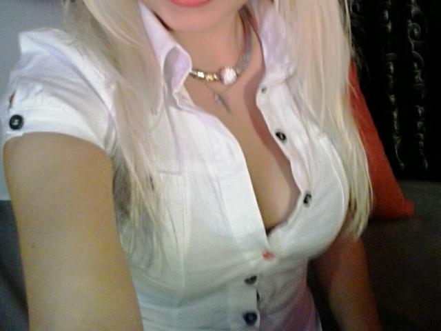 Leily, com cabelo loiro, camisa branca de botões e decote visível, mostrando seus seios grandes. Ela usa um colar de ouro e tem a camisa parcialmente desabotoada..