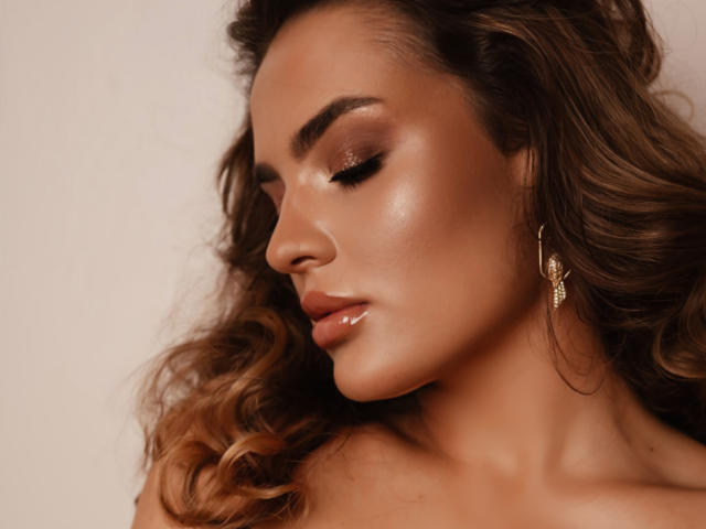 Celeste Belucci tiene labios marrones brillantes, piel radiante y cabello ondulado marrón. Lleva un arete dorado y tiene los ojos cerrados con sombra de ojos marrón brillante. Sus glúteos no son visibles..