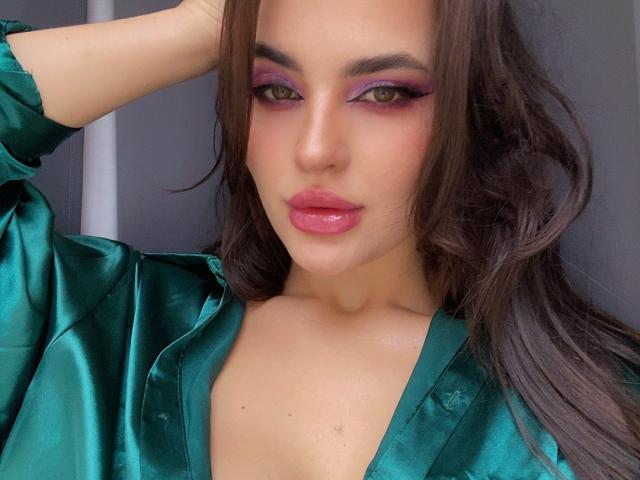 Celeste Belucci, met een glanzend groen zijden shirt, roze lippenstift en paars oogschaduw, heeft haar hand in haar bruine haar, staart met een serieuze uitdrukking en laat haar borsten zien..
