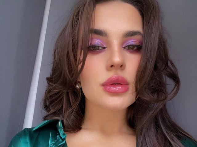 CelesteBelucci has glossy pink lips, purple eyeshadow, and wavy brown hair, wearing a green satin top, with a serious expression, looking directly at the camera.