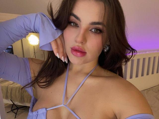 CelesteBelucci with long brown hair, wearing a purple, strappy top, exposing her tits, and purple earrings, has her hand on her head, looking seductively at the camera.