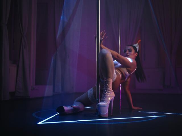Celeste Belucci, mit großen Brüsten und einem glänzenden silbernen Stange, sitzt auf einem dunklen Boden, trägt weiße Dessous und transparente Plateaustiefel, ihr Hintern ist angehoben und ihre Beine gespreizt..