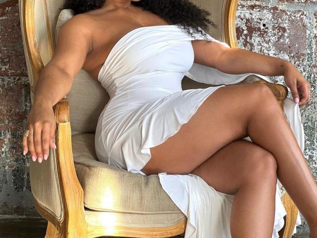 HotLolaXO con un grande sedere, seni nudi, in un vestito bianco, seduta su una sedia di legno, gambe incrociate, capelli ricci scuri, pelle abbronzata, anello alla mano sinistra, parete con texture..