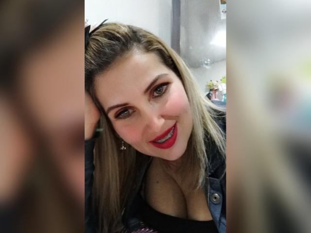 GiuliaAddams con capelli biondi, rossetto rosso e giacca di pelle nera, che mostra il suo seno prosperoso, sorride alla fotocamera, con una mano tra i capelli, una persona sfocata in primo piano..