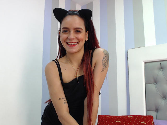 MaiaPark with red hair, black cat ears, smiling, black dress, cross necklace, tattoo on right arm, sitting on red pillow, bed with white frame in background.