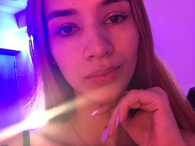 GretaHarris avec un éclairage néon rose-violet sur le visage, de longs cheveux blonds, un maquillage subtil, des lèvres roses, des ongles blancs, tenant son menton, regardant la caméra, avec un léger sourire..