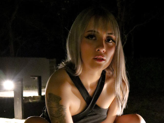 SabrinaCole, con cabello rubio, camiseta negra ajustada y tatuaje en el brazo derecho, agachada en una luz tenue, mirando a la cámara con labios rojos, luz suave desde la izquierda, ambiente nocturno..