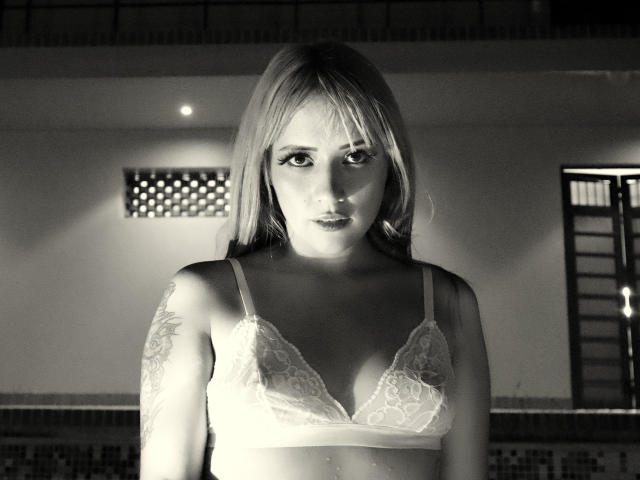 Fotografia in bianco e nero di SabrinaCole con lunghi capelli biondi, che indossa un reggiseno in pizzo, mettendo in mostra il suo seno piccolo e un tatuaggio sul braccio destro, mentre guarda intensamente la fotocamera..