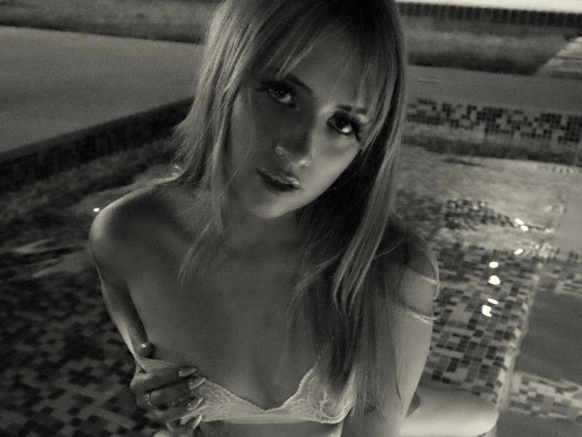 Foto em preto e branco de SabrinaCole com cabelo comprido, olhando para cima, usando um sutiã de renda, seus pequenos seios levemente visíveis, em pé ao lado de uma piscina com azulejos, com as mãos perto do peito..