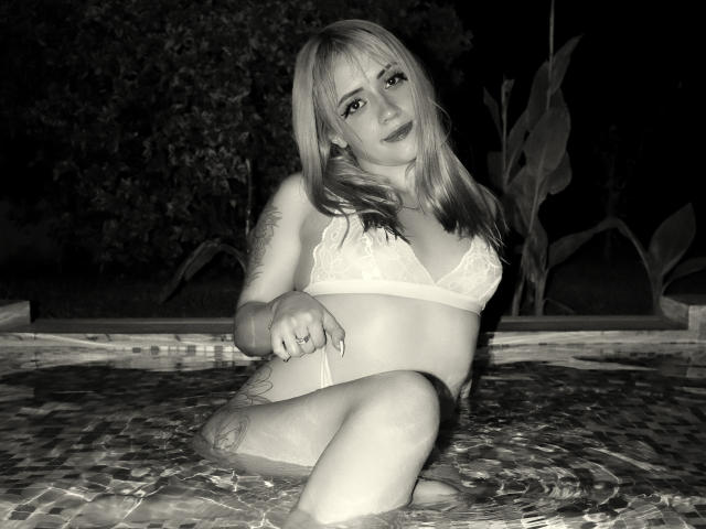 SabrinaCole, avec ses cheveux blonds, son soutien-gorge en dentelle et sa culotte, assise au bord d'une piscine la nuit, montrant des tatouages et une bague, les jambes croisées, regardant la caméra..
