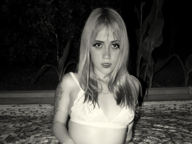 Black-and-white photo of SabrinaCole with long blonde hair, wearing a lace bra, standing in a tiled pool, looking directly at the camera, showing a tattoo on her right arm.