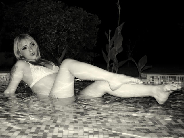 SabrinaCole está en una piscina, vestida con un sostén y tanga blancos de encaje, con las piernas cruzadas, mostrando sus pechos y vulva, en blanco y negro, de noche, en una pose relajada..