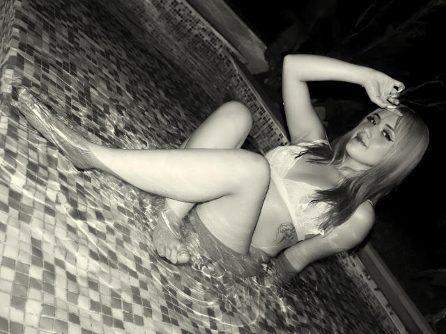 Photo en noir et blanc de SabrinaCole dans une piscine, portant un bikini en dentelle, les jambes relevées, une main dans les cheveux, de l'eau sur sa peau, un tatouage sur le côté droit, regardant l'appareil photo..
