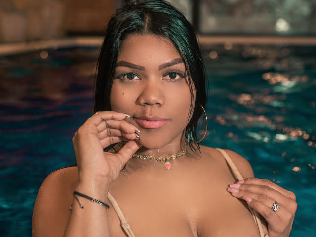 NickyBernad mit nassen, dunklen Haaren, brauner Haut, Reifenohrringen, einer rosa Halskette und pinkem Lippenstift, die ihre Wange in einem Schwimmbecken berührt, wobei ihre Brüste und ein Ring an ihrer rechten Hand zu sehen sind..