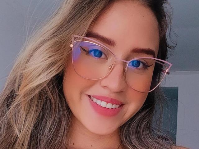 KylieSaoko has blue eyes, pink glasses, and a smile with pink lipstick, showing off her cute face and blonde wavy hair. She's looking directly at the camera.