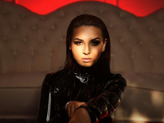 KayaOcean with dark, slicked-back hair, wearing a shiny black leather jacket, sitting with arms crossed, serious expression, red lighting, and a tufted gray couch behind her.