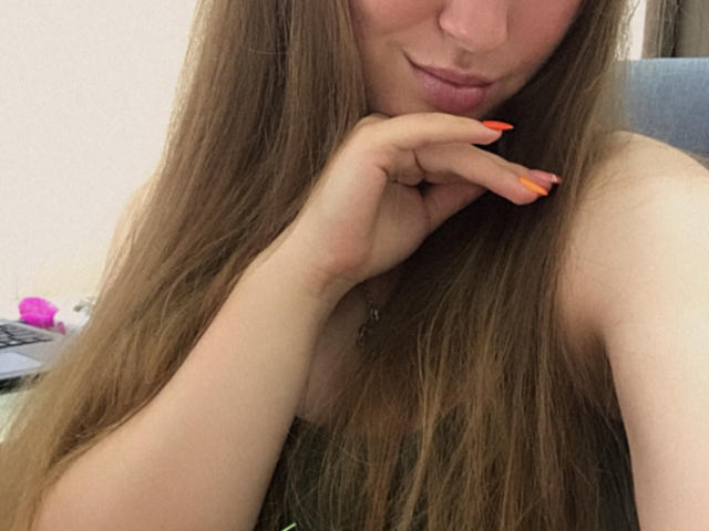 Dooris with long brown hair, pouty pink lips, and orange nails, touching her chin, showing her bare shoulders and a hint of her small tits.