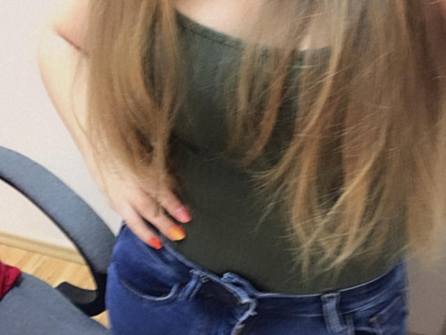 Doris, avec de longs cheveux blonds, un débardeur vert et un jean bleu, une main sur la hanche, des ongles peints en orange et rose, la partie inférieure de sa poitrine visible, sans visage..
