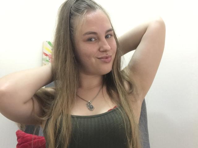 Een vrouw met lang blond haar, een groene top, armen omhoog, lachend, kleine borsten, een ketting, lichte huid, een roze kussen op de achtergrond, handen in haar haar, een zachte uitdrukking, een nonchalante pose..