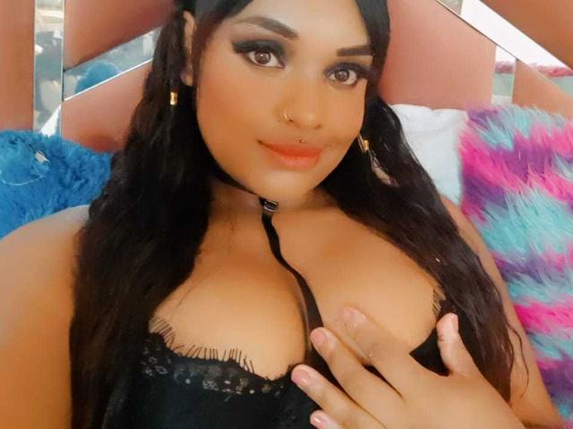AllyCharlotee met lang zwart haar, bruine ogen en een neusring, draagt een zwarte kanten bh, heeft haar hand op haar borsten, een choker en gouden oorbellen, en glimlacht naar de camera..