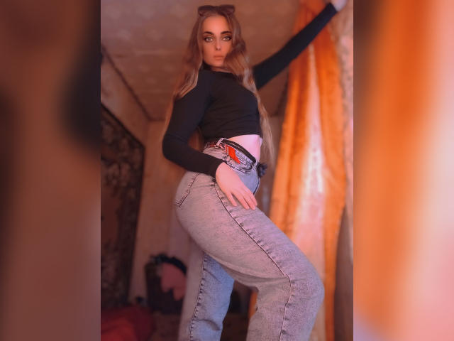 KariMillie, avec ses longs cheveux blonds, son haut noir et son pantalon gris, debout les fesses en l'air, regardant par-dessus son épaule, dans une pièce faiblement éclairée..