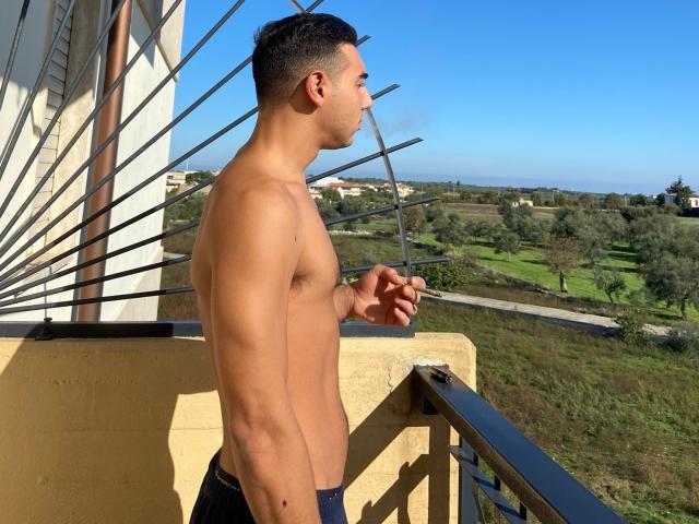 GiovaneBello, shirtless, standing on a balcony, holding a cigarette, blue jeans, muscular, tan skin, short dark hair, clear blue sky, metal railing, yellow wall, green landscape, sunlit.