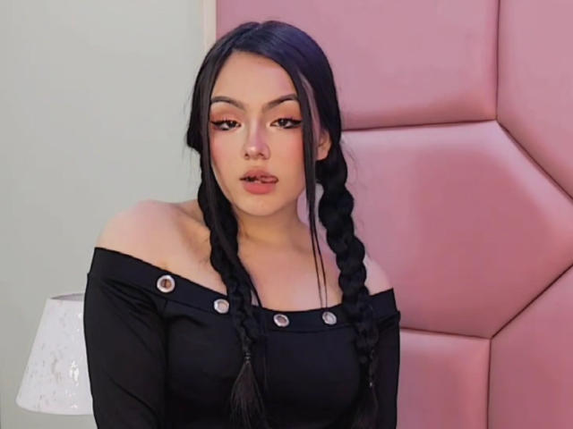 HaleiBeckham con trenzas negras, blusa negra con un hombro descubierto, cuello con botones, pared rosa y cabecero rosa. Tiene un rostro sexy y lindo con maquillaje rosa en los ojos..