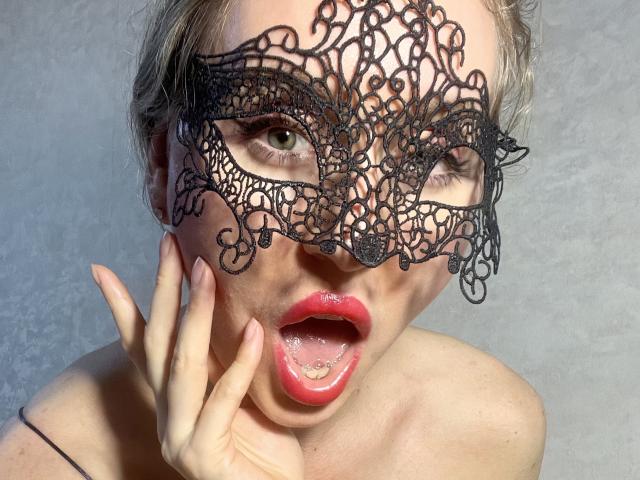 TheHottestBlonde with a black lace mask, open mouth showing red lipstick, hand near face, blonde hair, pale skin, green eyes, and no clothes, looking sexy and mysterious.
