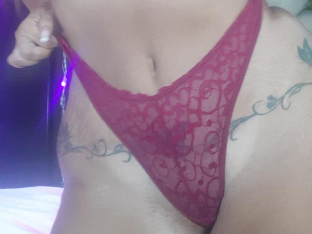 DanielaXHotty's red lace thong is pulled up, showing her tattooed hips and a small, visible part of her pussy. Her hand is holding the side.