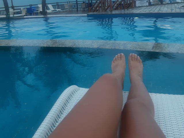 DanielaXHotty's legs with tanned skin, feet in the blue pool, resting on a white wicker chair, water reflection, poolside setting, daytime.