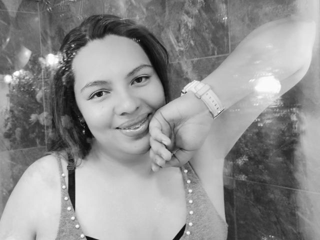 YinethBeilly, con el torso visible, sonriendo, vistiendo una camiseta sin mangas con tachuelas, un reloj de pulsera y aretes, mirando a la cámara, en blanco y negro, con su mano cerca de su boca..