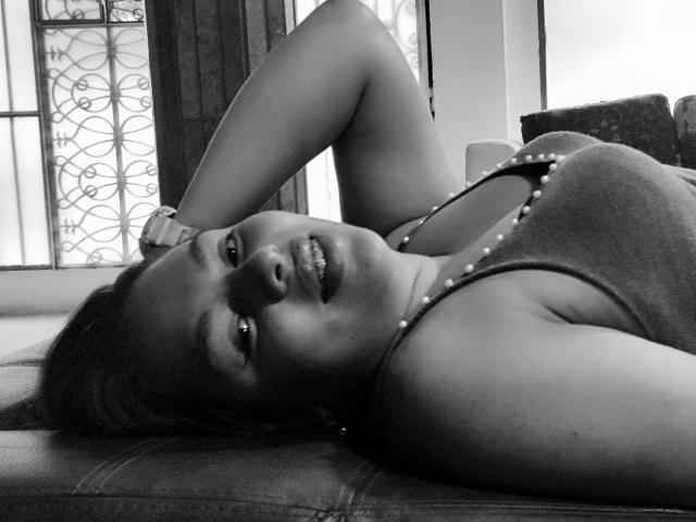 YinethBeilly acostada en un sofá, con el brazo detrás de la cabeza, sonriendo, vestida con una camiseta sin mangas con tachuelas, en blanco y negro, mostrando sus pechos y su linda cara, relajada, mirando a la cámara..