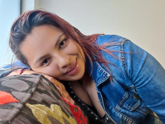 YinethBeilly with red-dyed hair, smiling, wearing a blue denim jacket, resting her head on a colorful pillow, showing her cleavage. Her brown eyes and cute nose are visible.