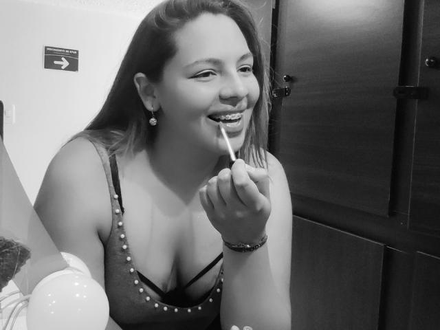 YinethBeilly, con senos de tamaño medio, sonriendo, sosteniendo un caramelo en la boca, vistiendo una blusa adornada con cuentas, en una foto en blanco y negro, con cabello largo y aretes..