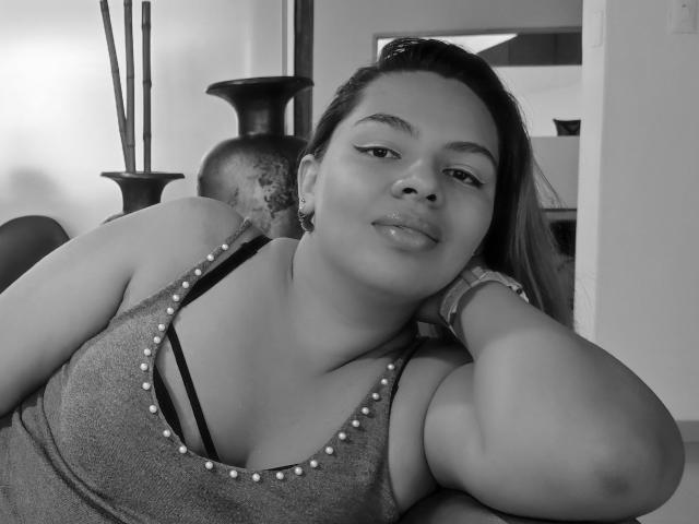 YinethBeilly, avec des lèvres pulpeuses, portant un débardeur scintillant, montrant ses seins, avec un bandeau de soutien-gorge noir visible, appuyée sur son bras, regardant la caméra, photo en noir et blanc, détendue..