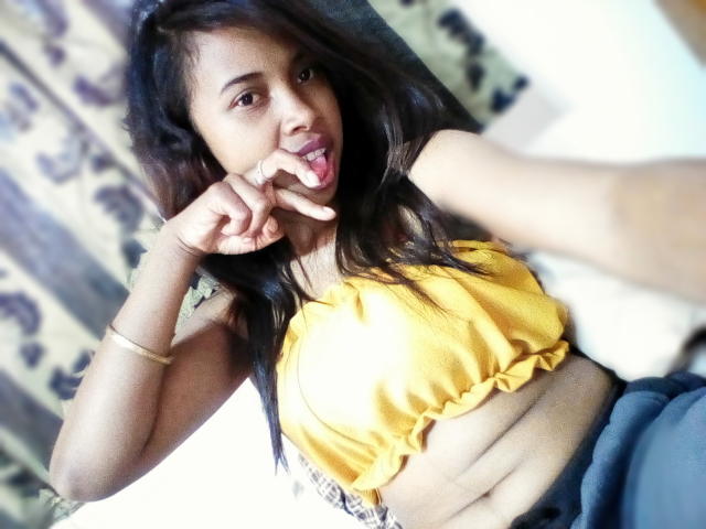Ckamii, con cabello oscuro, un top corto amarillo y la lengua afuera, haciendo un gesto de "shh", mostrando sus pequeños pechos y abdomen. Lleva pantalones azules y una pulsera dorada..