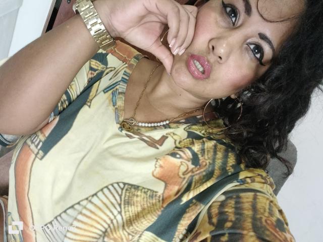 YiyaHot69 with curly black hair, gold wristwatch, hoop earrings, and pink lipstick, wearing an Egyptian-themed top, touching her face with a surprised expression.