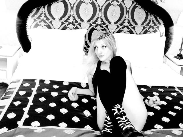 Fotografia in bianco e nero di MissCherryOnTop con lunghi capelli biondi, seduta su un letto, che indossa calze a righe zebrate fino alla coscia, una maglietta nera e nient'altro, mostrando il suo sedere scoperto..