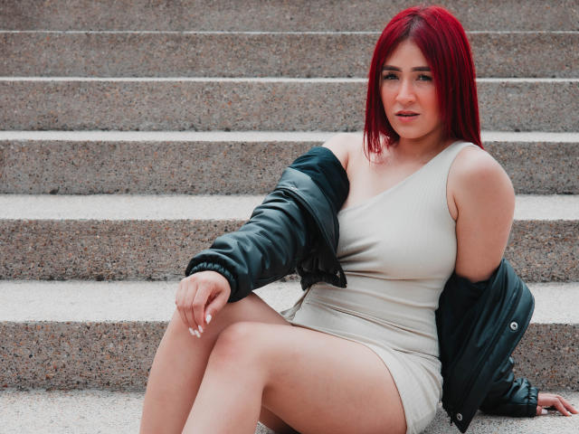 Nico Coleman, mit roten Haaren, heller Haut und einer kurvigen Figur, sitzt auf steinernen Stufen und trägt ein beiges Kleid und eine schwarze Lederjacke, wobei ihr Hintern freiliegt und ihre Brüste sichtbar sind..