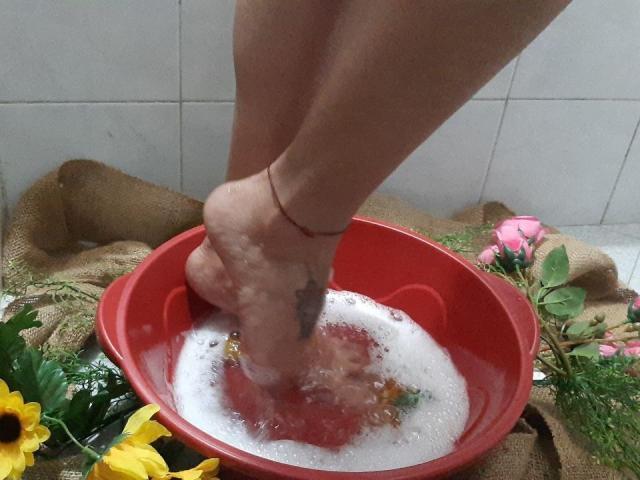 El pie de Laila Castillo, con un pequeño tatuaje, un brazalete en el tobillo y un cordón rojo, está lavando un cuenco rojo lleno de agua jabonosa y rosas rosadas..