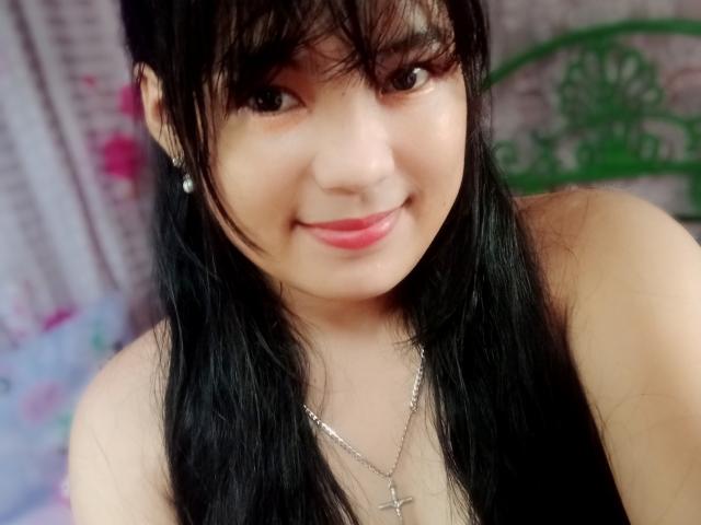 SweetAsian with black hair, bangs, and pink lips, smiling softly. She's wearing a cross necklace, has small earrings, and is showing her bare shoulders.